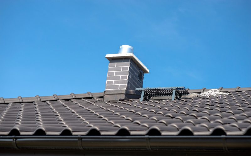 The roof of a single-family house covered with a new ceramic tile in anthracite against the blue sky. Vsible system chimney covered with tiles.